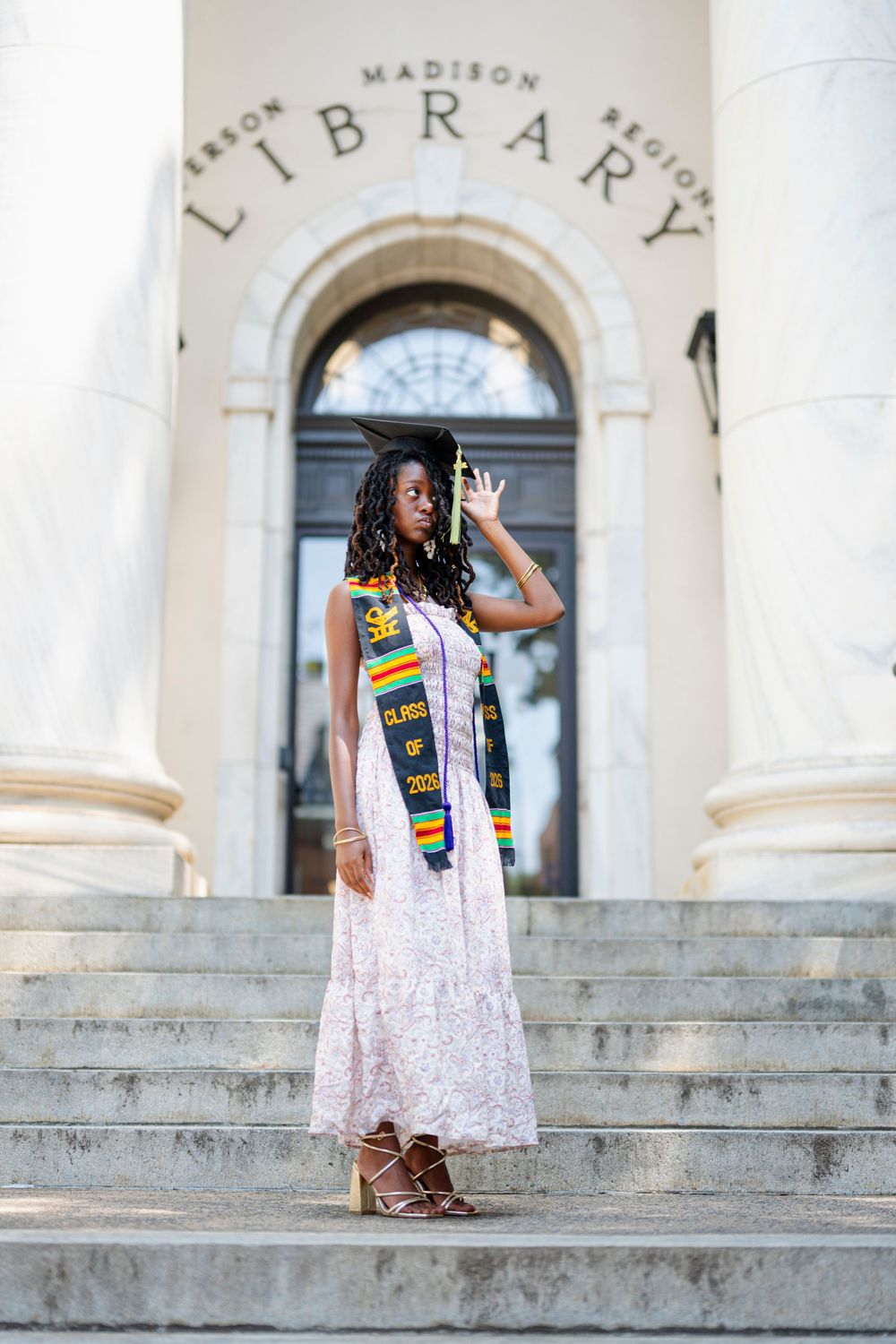 A graduate in a floral dress and kente stole poses proudly on library steps holding her cap.