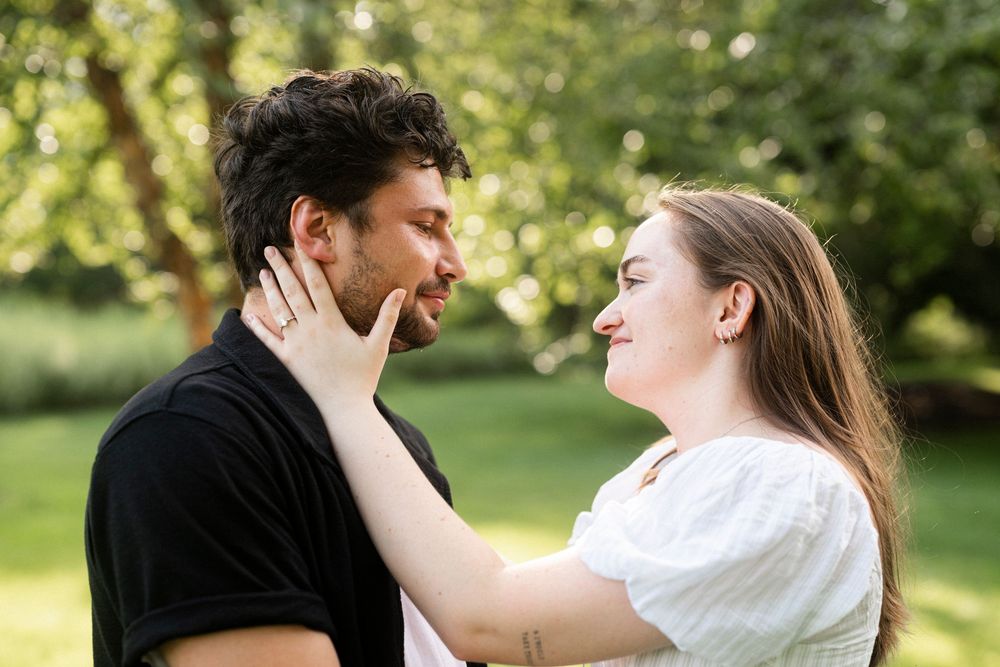 Couple sharing a romantic moment outdoors, woman smiling and touching man's face in a lush green park.