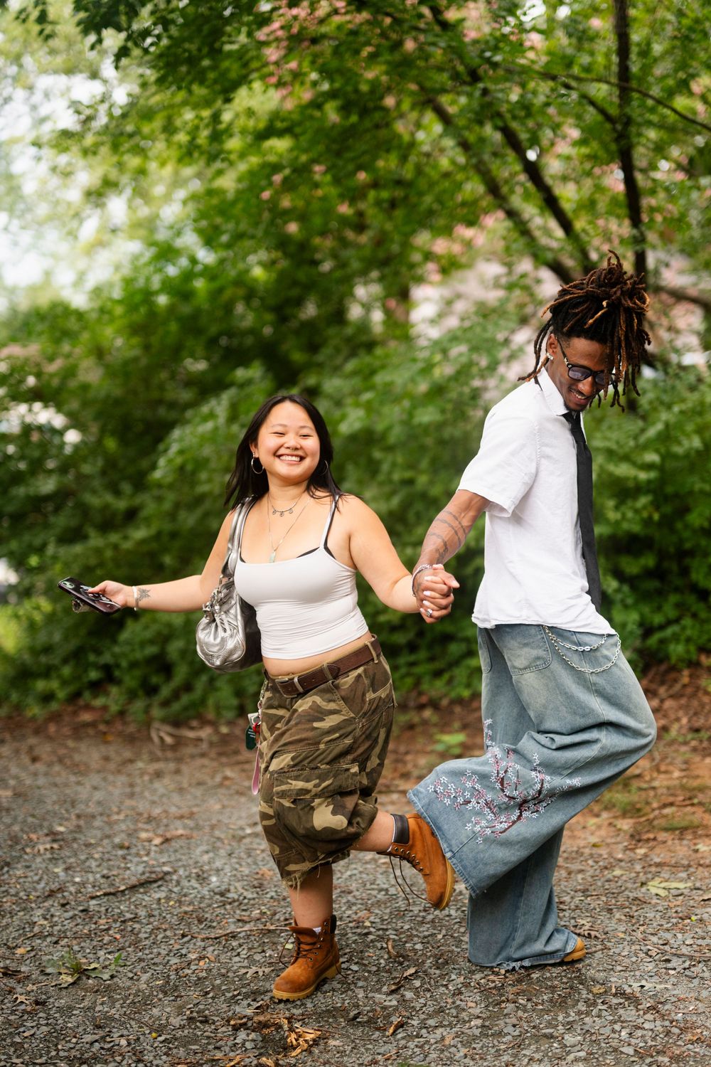 Happy couple holding hands and dancing outdoors, woman in camo skirt and man with dreadlocks, surrounded by lush green trees.