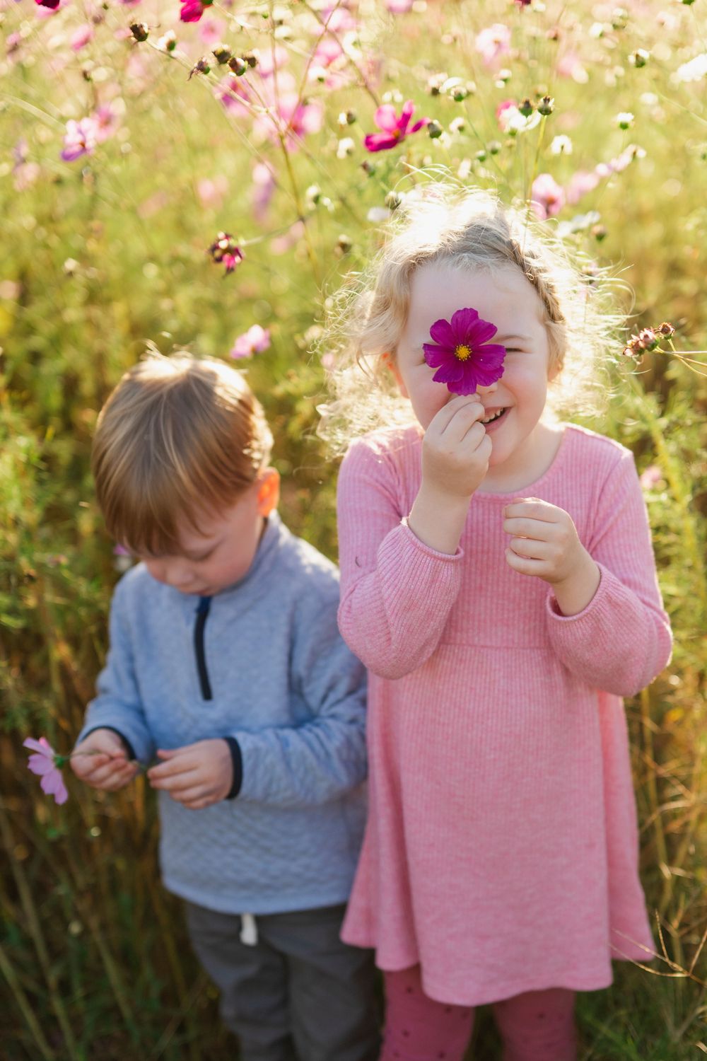 Two young children play in a colorful wildflower meadow, girl in pink dress holding flower over her eye.