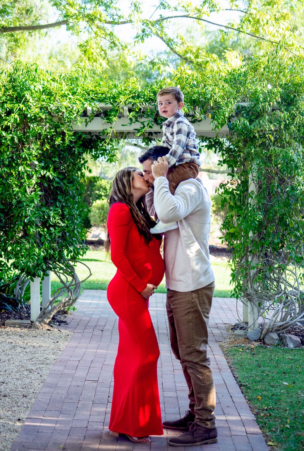 Romantic maternity photoshoot with couple in red dress and casual attire on brick path.