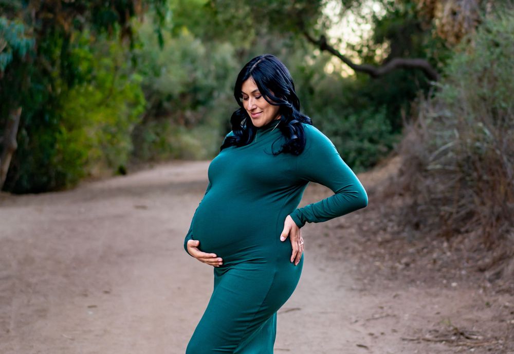 A pregnant woman in an emerald green dress poses on a nature trail with sunlit trees in the background.