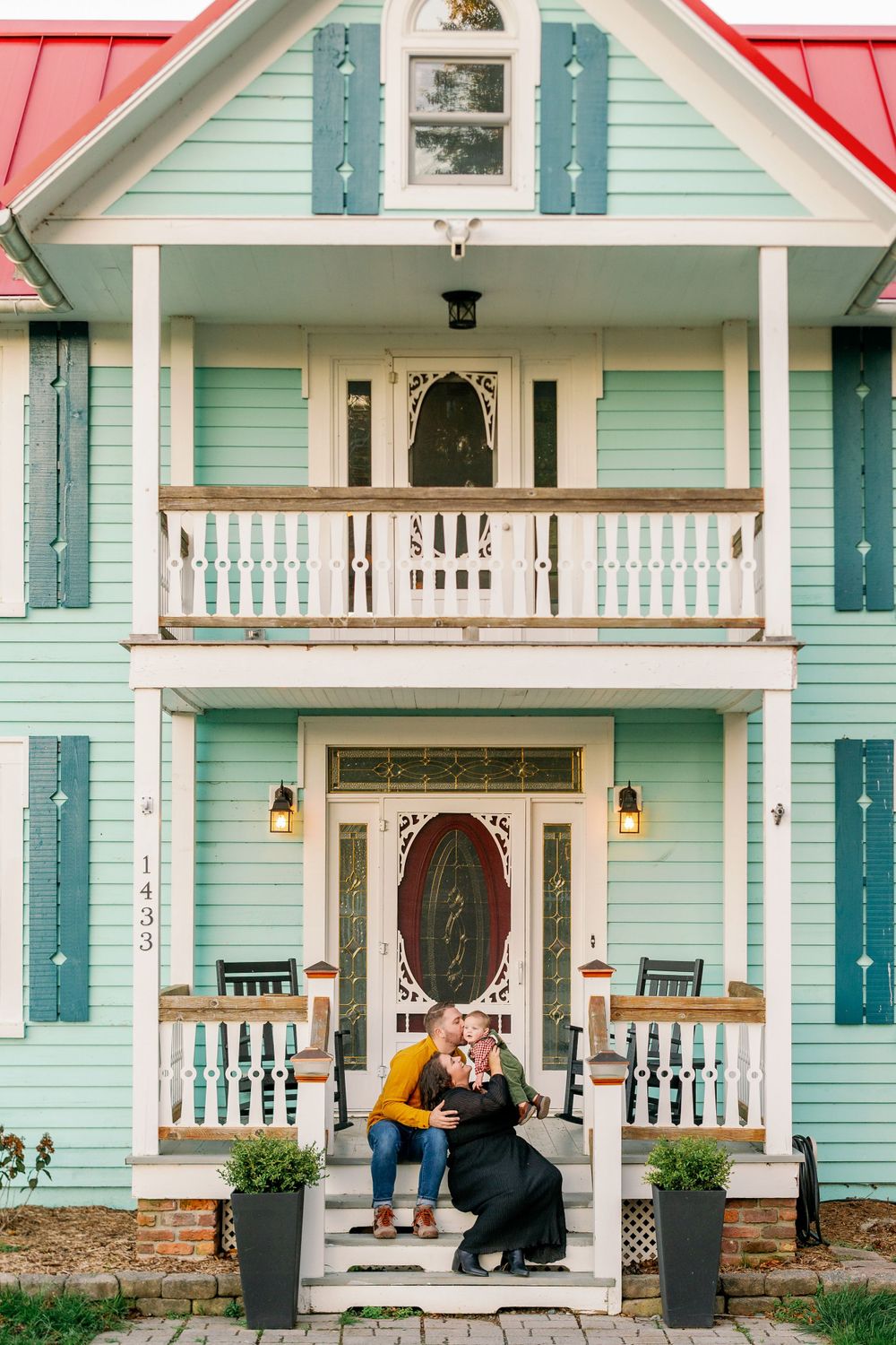 A couple sitting on the front porch steps of a mint green Victorian-style house.