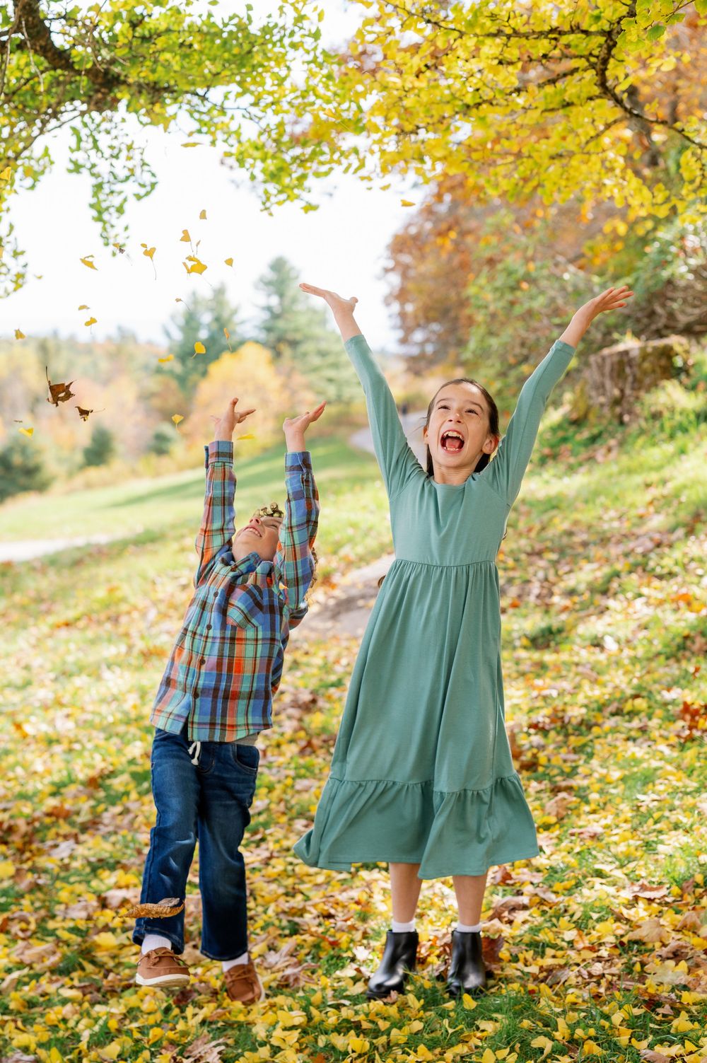 Two children joyfully tossing autumn leaves in a colorful fall park, wearing seasonal outfits.