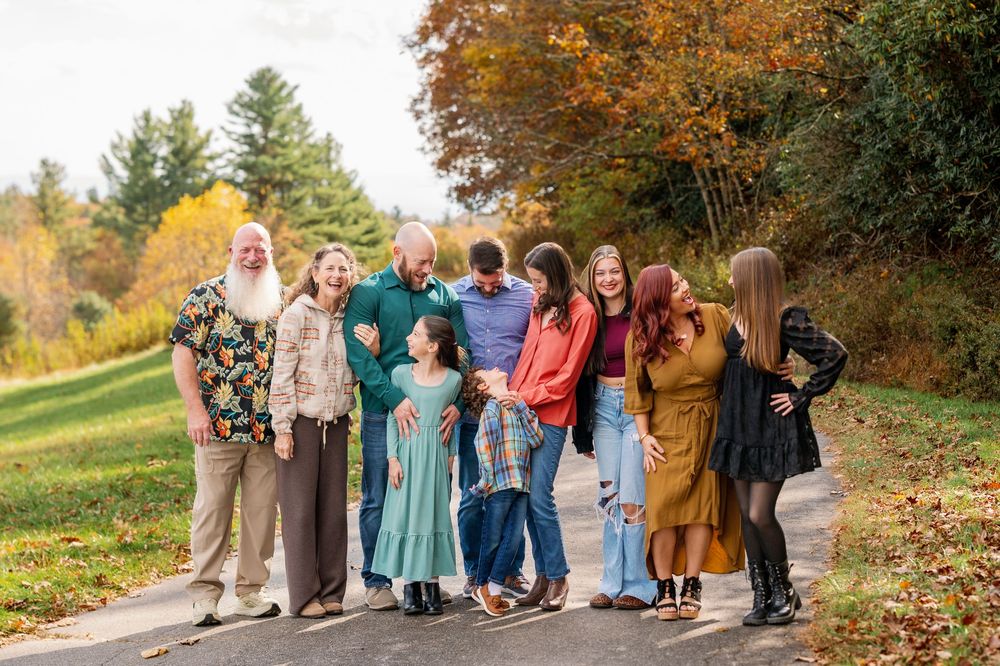 Large family group photo on a tree-lined path during autumn, wearing colorful outfits in fall foliage setting.
