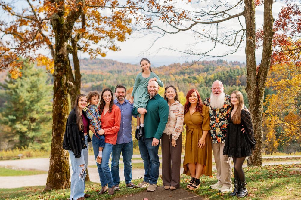 Large family group portrait outdoors in autumn, smiling together among colorful fall foliage and scenic mountain views.