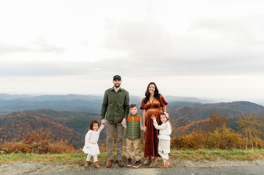Family of five posing on mountain overlook with fall foliage backdrop during autumn outdoor photo session.