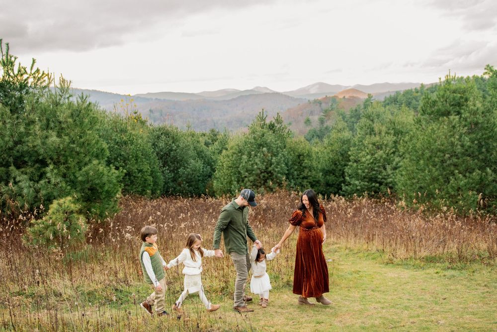 Family of five holding hands walking through a meadow with mountain views and lush green trees in the background.