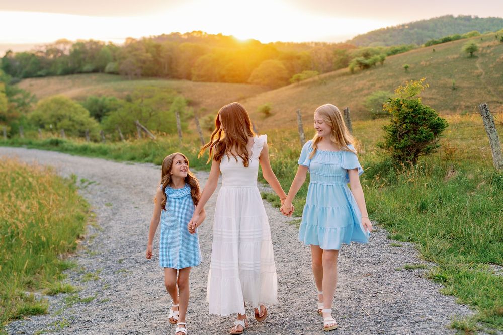 Three girls in blue and white dresses walk hand-in-hand down a gravel path at golden sunset.