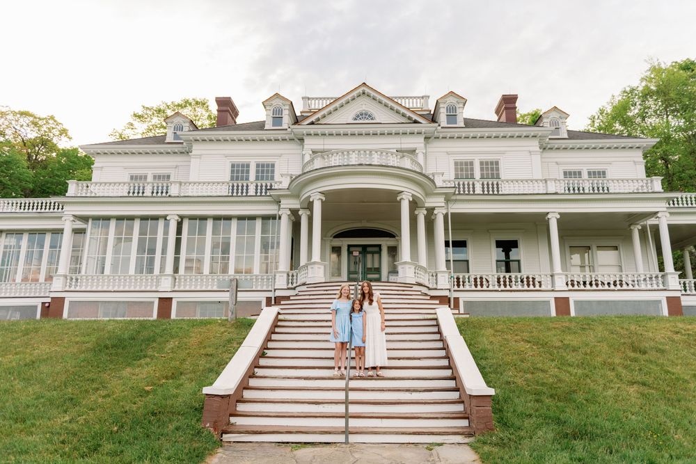 Two women pose on grand steps of a white colonial mansion with wraparound porch and brick accents.