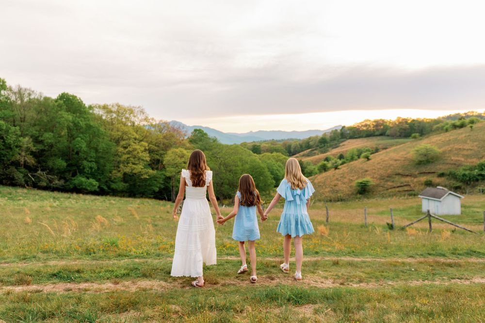 A mother and two daughters in blue and white dresses hold hands overlooking rolling green hills at sunset.