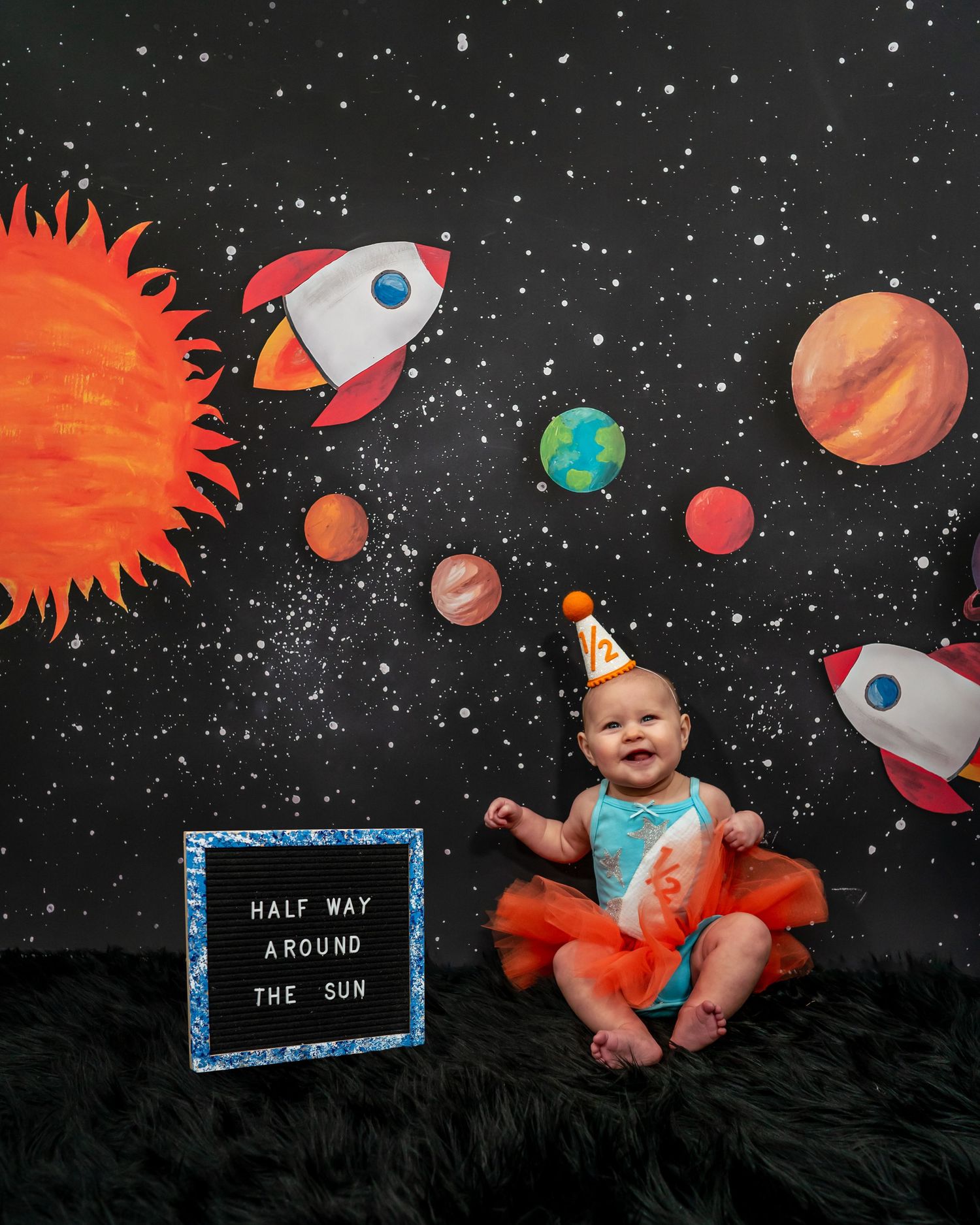 Joyful baby in orange tutu celebrating half birthday with space-themed backdrop and rocket ship decorations.