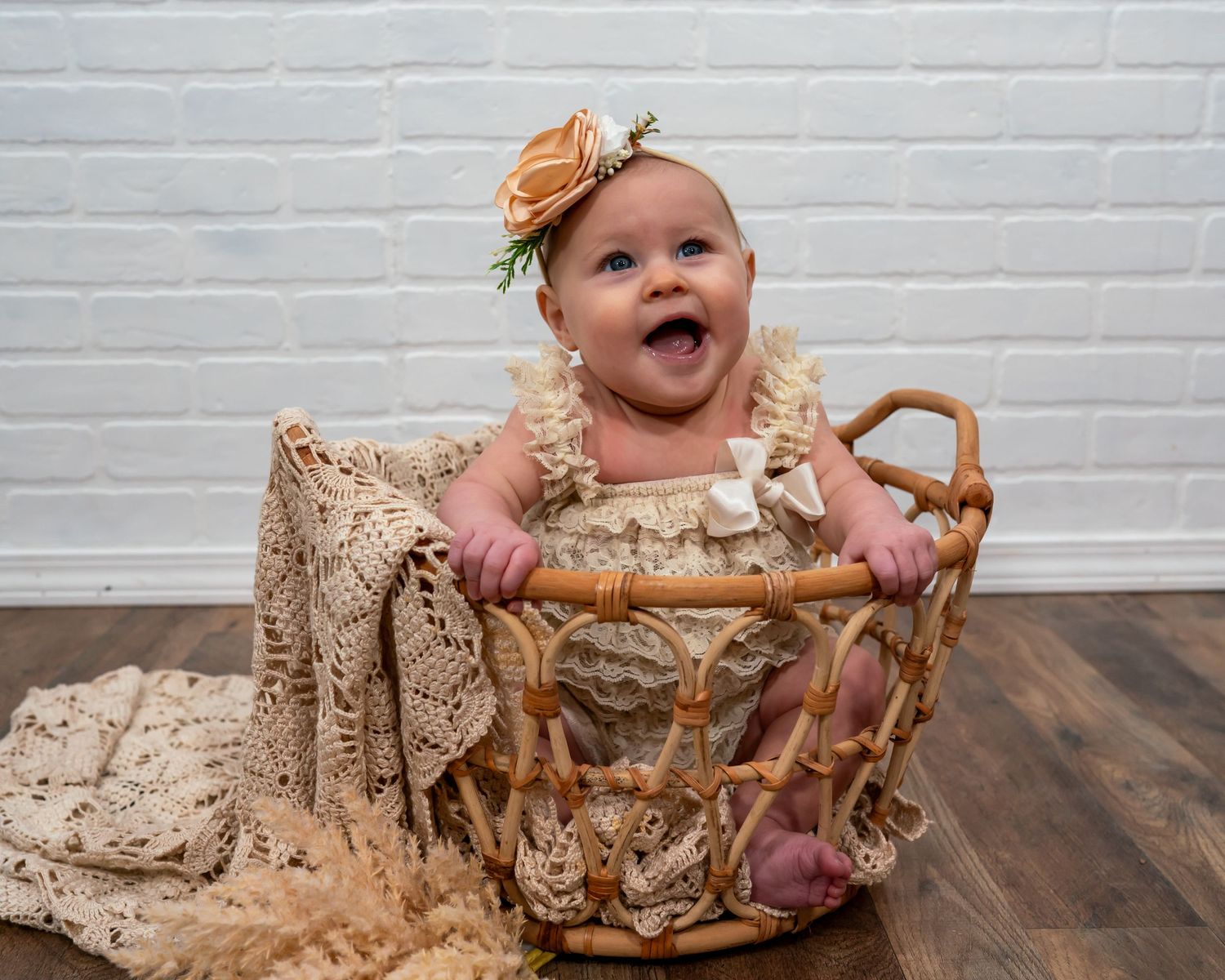 Happy baby girl laughing in a wicker basket wearing a lace romper and peach floral headband.