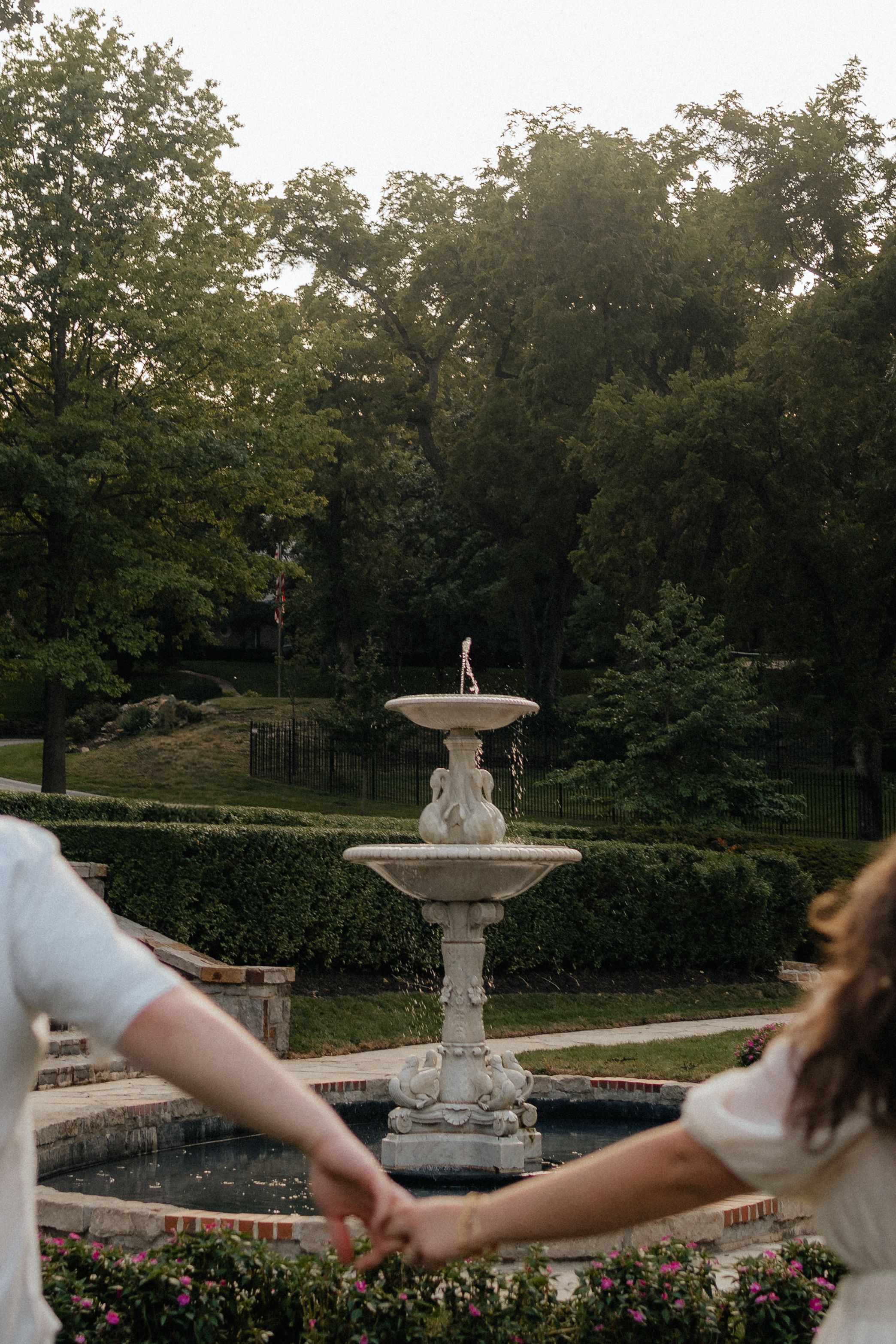 Stone fountain stands elegantly in a formal garden with pathways and lush trees at dusk.