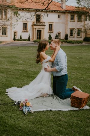 A couple shares an intimate moment on a blanket in front of a stately mansion.