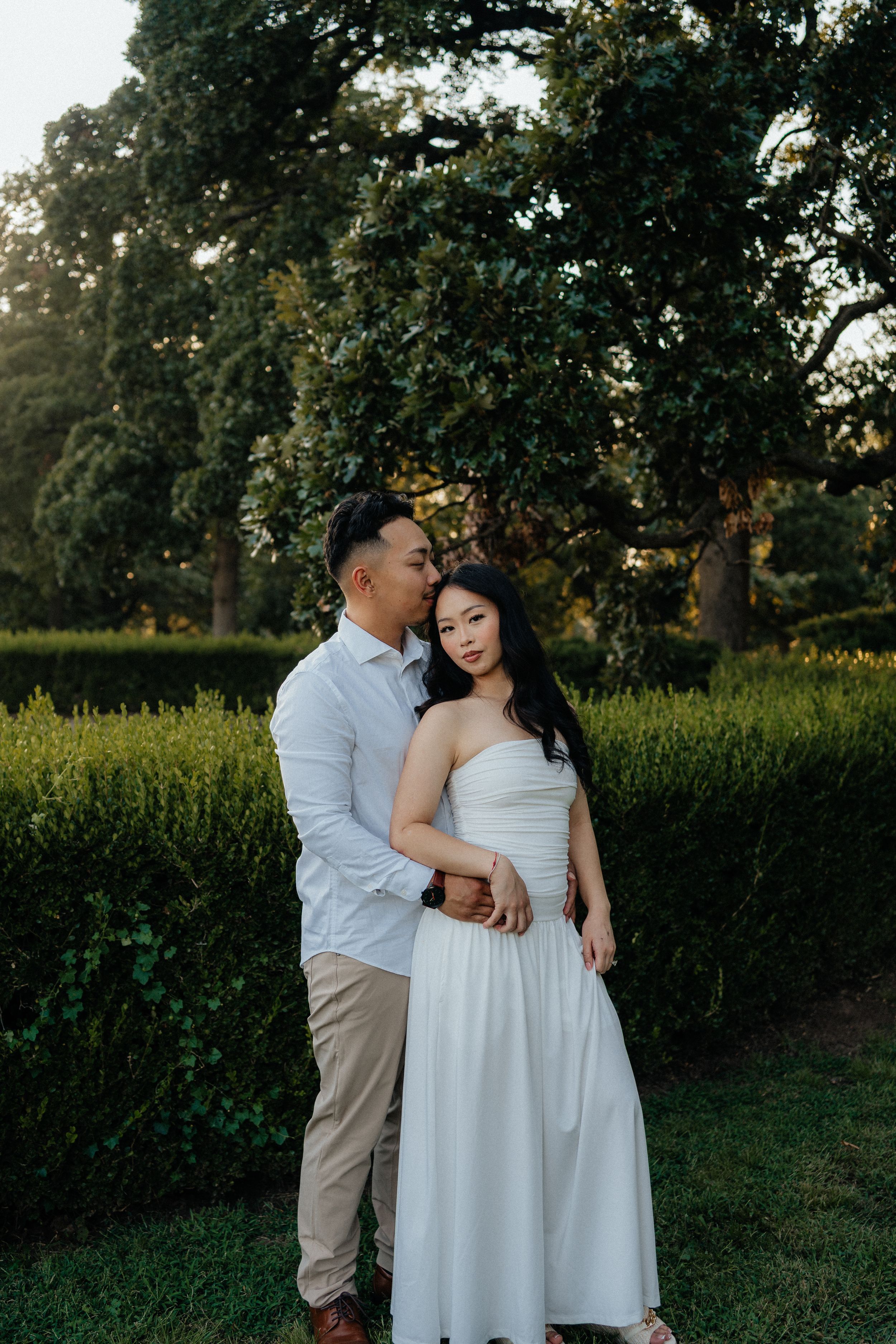 Romantic couple embraces in a garden at sunset with lush trees and greenery in the background.