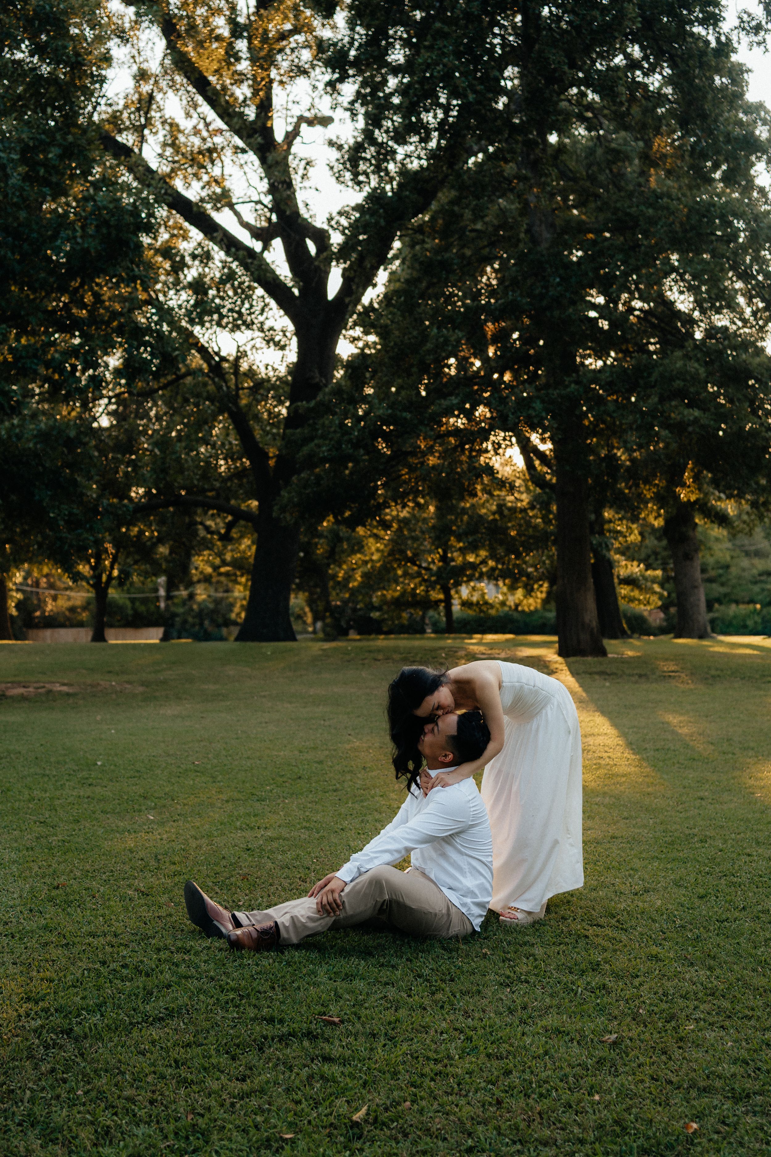 A couple in white outfits reclines on grass in a sunlit park during golden hour.