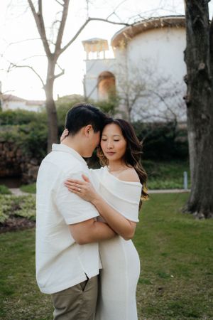 A couple in matching white outfits embraces during an outdoor engagement session near a historic building.