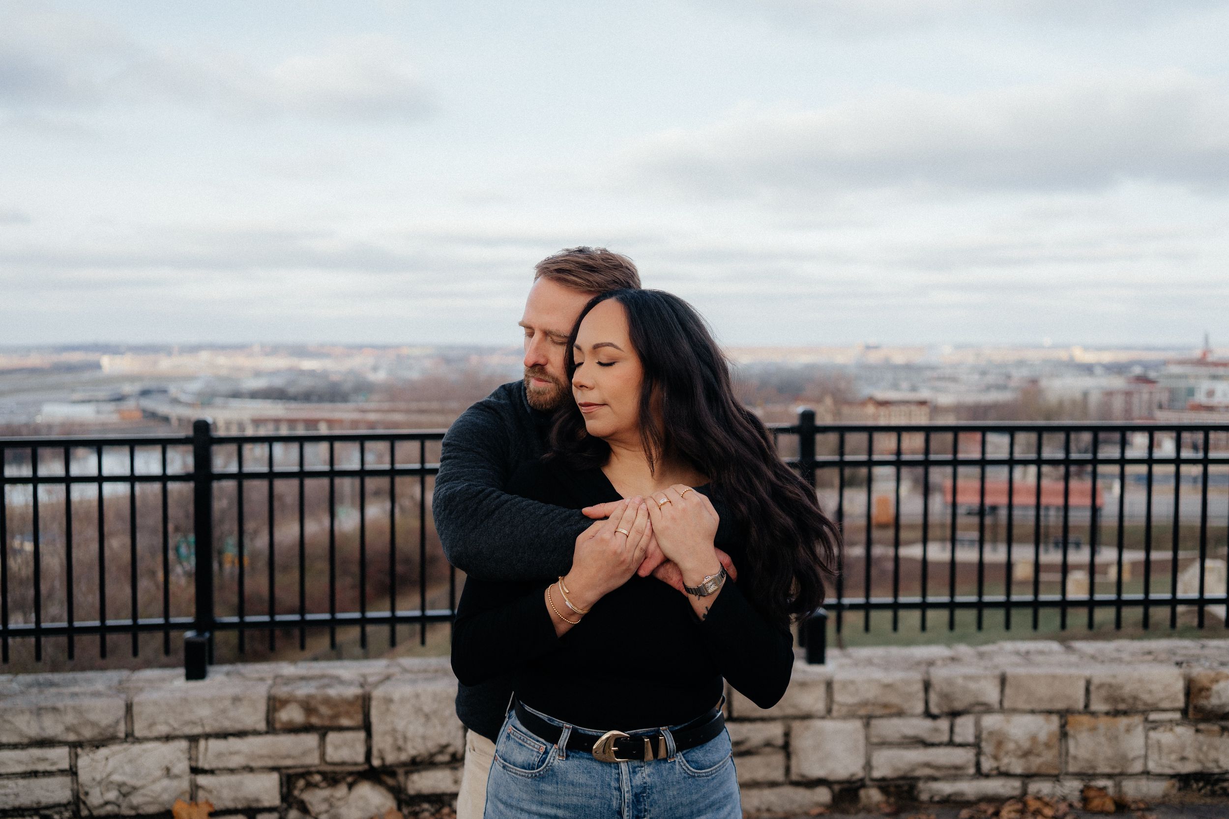 A couple embraces on a scenic overlook with stone walls and a cityscape view in the background.