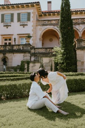 A romantic couple in white attire shares an intimate moment on manicured grounds of a Mediterranean villa.