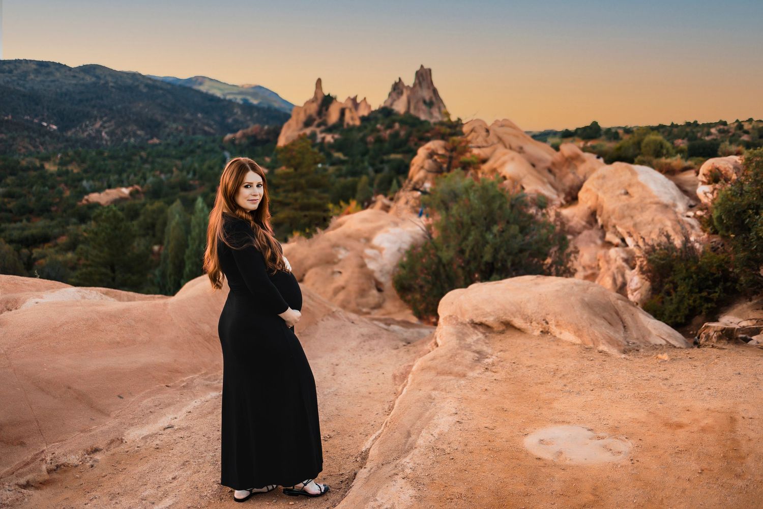 pregnant woman in long black dress, looking back at camera, cradling bump.  Maternity photoshoot at garden of the gods in colorado