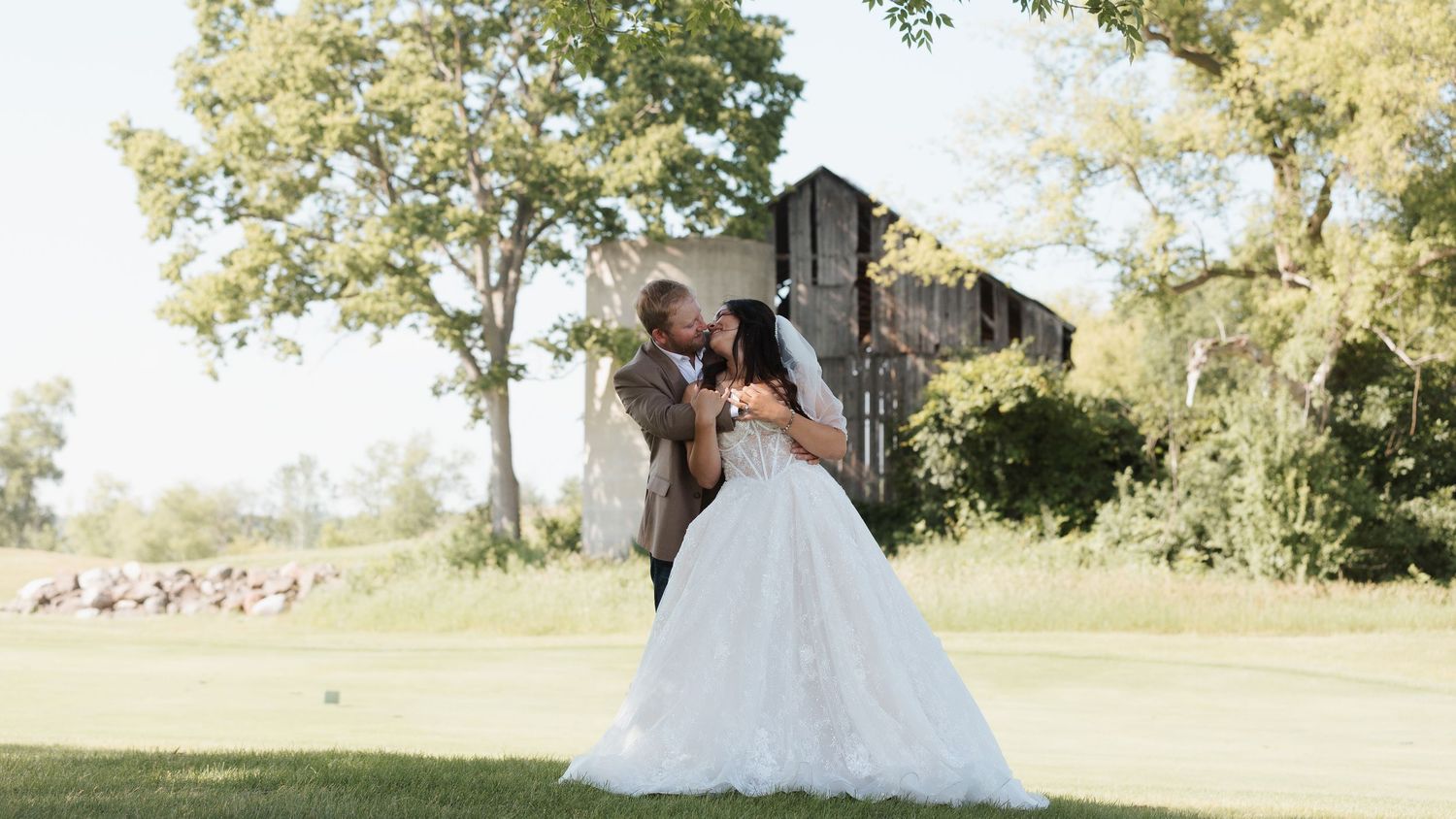 Bride and groom share a romantic kiss in a lush green outdoor wedding venue with a rustic barn backdrop.