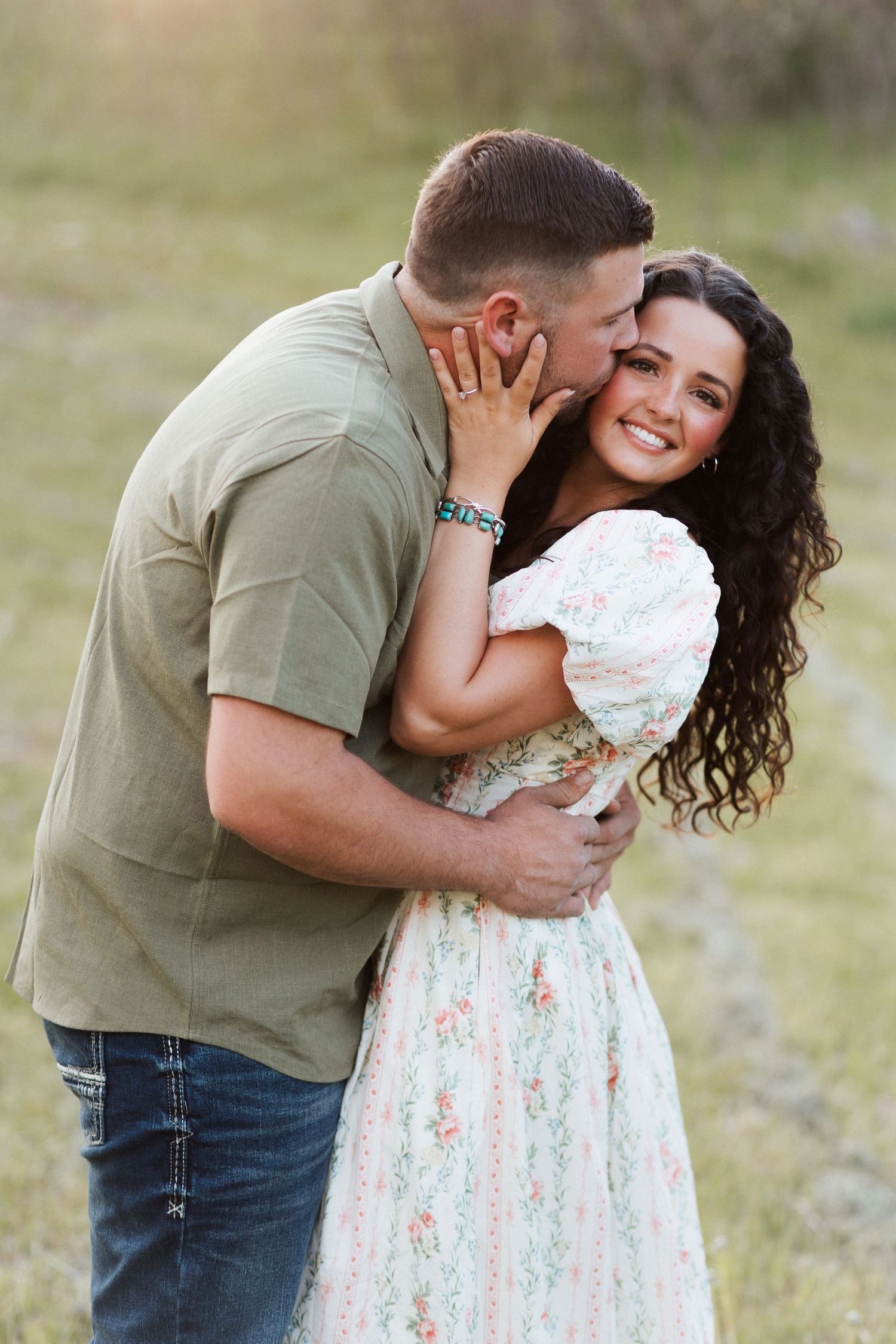 A couple embraces outdoors, man in green shirt kisses smiling woman in floral dress with curly dark hair in golden light.