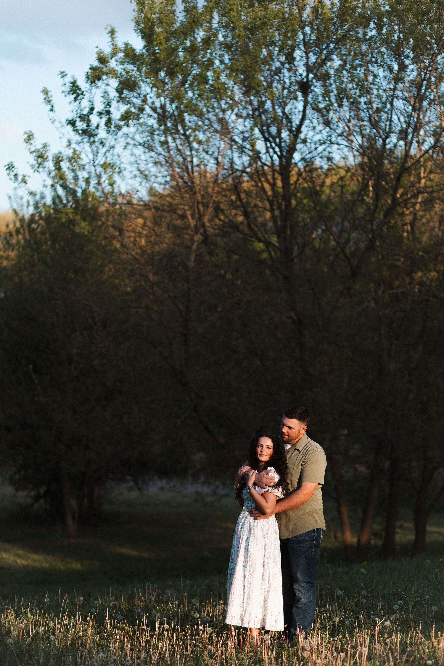 Couple embracing in a sunlit field surrounded by lush green trees during golden hour.