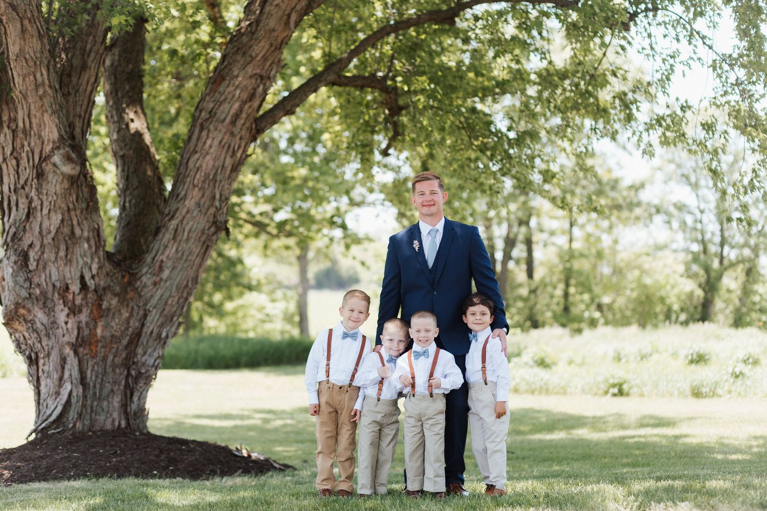A groom in a navy suit poses with four young ring bearers in suspenders under a large tree outdoors.