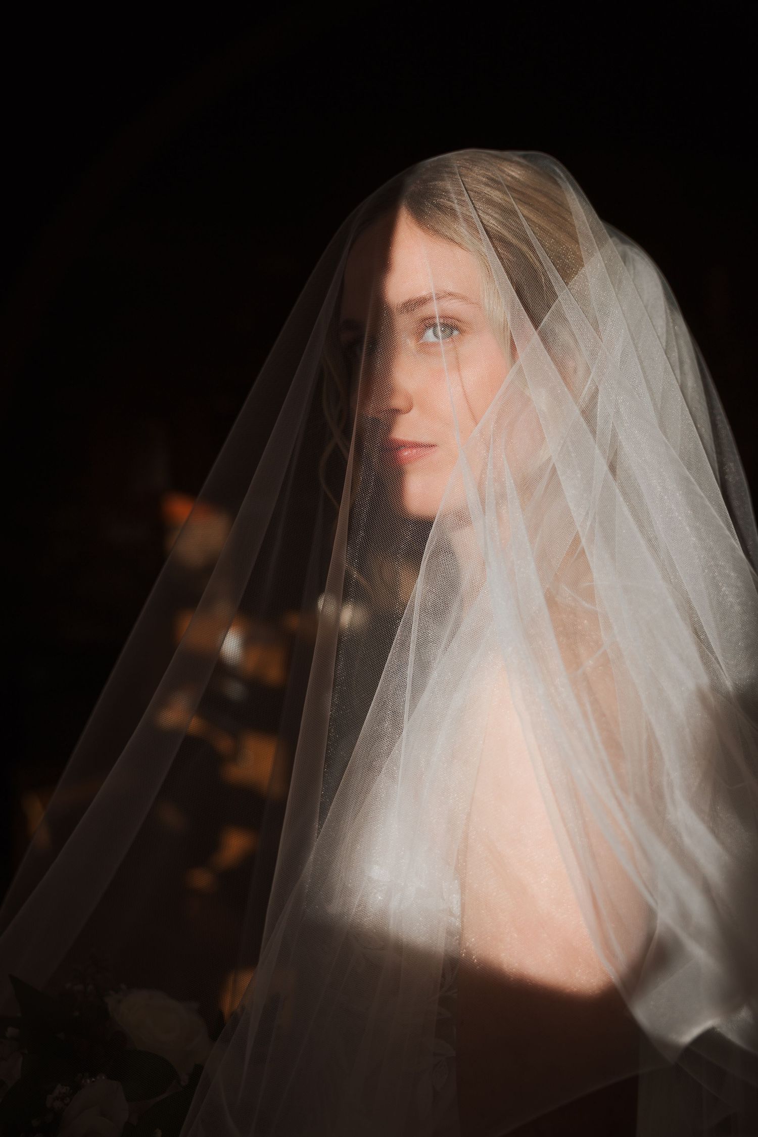 Dramatic side profile of bride wearing delicate white veil against dark background.