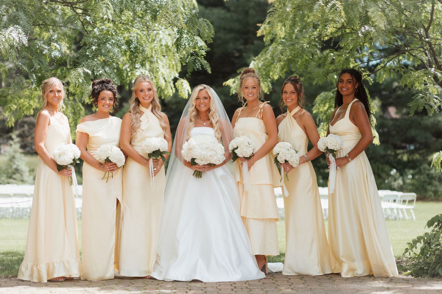 Bride in white gown poses with six bridesmaids in cream dresses holding white bouquets outdoors.