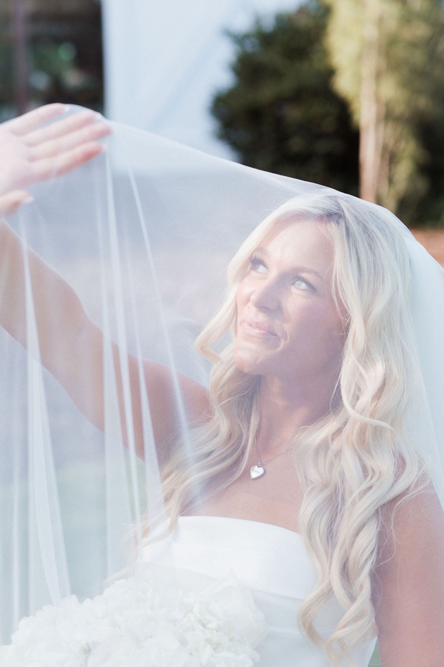 Smiling blonde bride lifting her white veil outdoors, wearing a strapless gown and heart necklace on her wedding day.