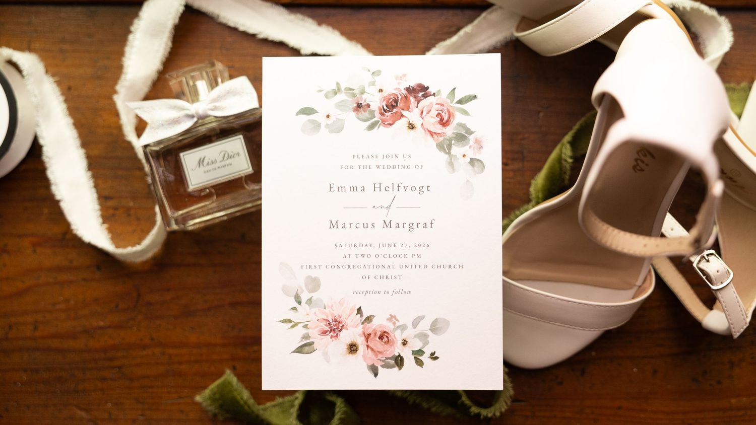 Elegant wedding invitation with floral design beside white heels and Dior perfume on wooden surface.