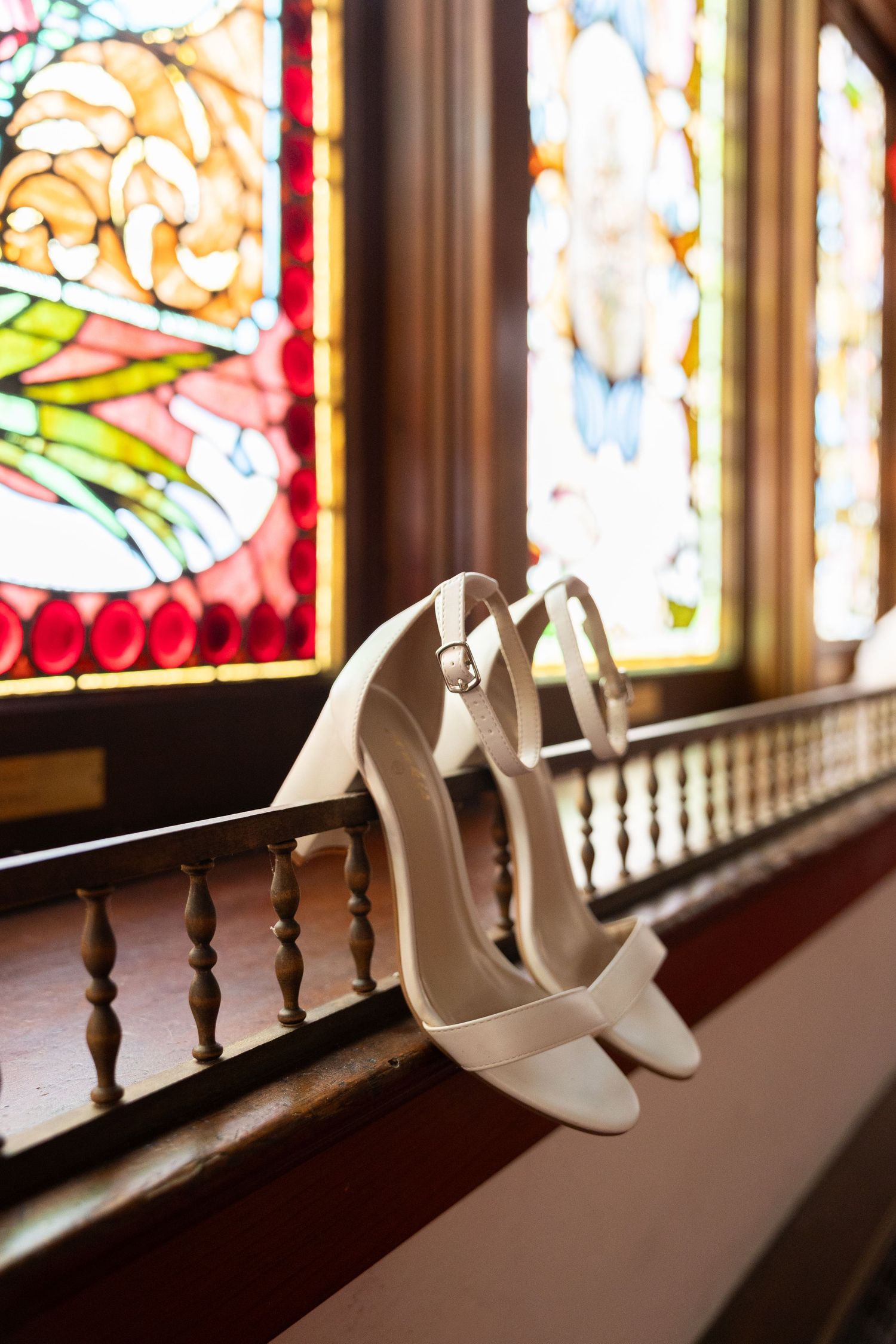 White bridal heels resting on a wooden railing in front of colorful stained glass windows in a church.