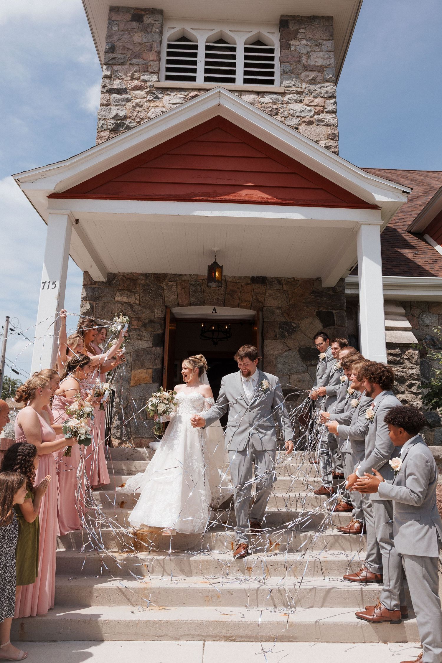 Bride and groom exit stone church on wedding day as guests celebrate with streamers on sunny steps.