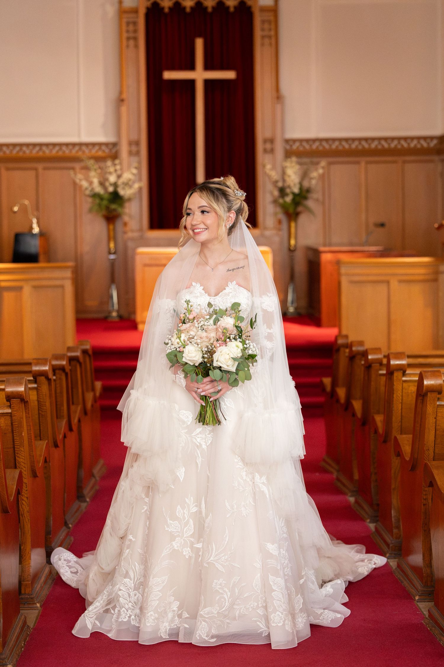 Bride in lace wedding gown and veil holds bouquet at church altar with wooden pews and cross backdrop.