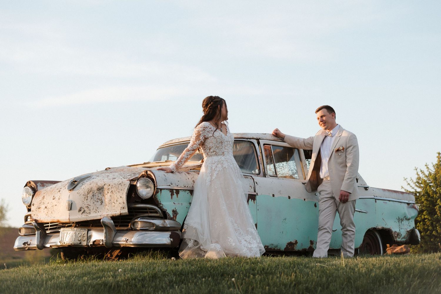 Bride and groom posing beside a rustic vintage teal car on a green lawn at golden hour.