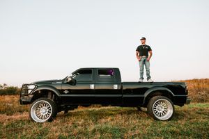 A black Ford F-250 pickup truck with custom wheels parked on a grassy hill during sunset.