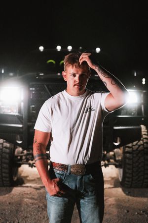 A person in a white t-shirt and jeans stands in a dimly lit outdoor setting with dramatic lighting effects.
