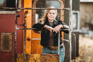 A person in denim leans against a rustic orange and red train car while posing for a portrait.