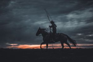 A silhouetted figure rides a horse against a dramatic stormy sky at sunset.