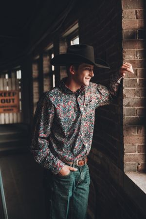 Candid portrait of someone in western wear and cowboy hat leaning against a brick wall.