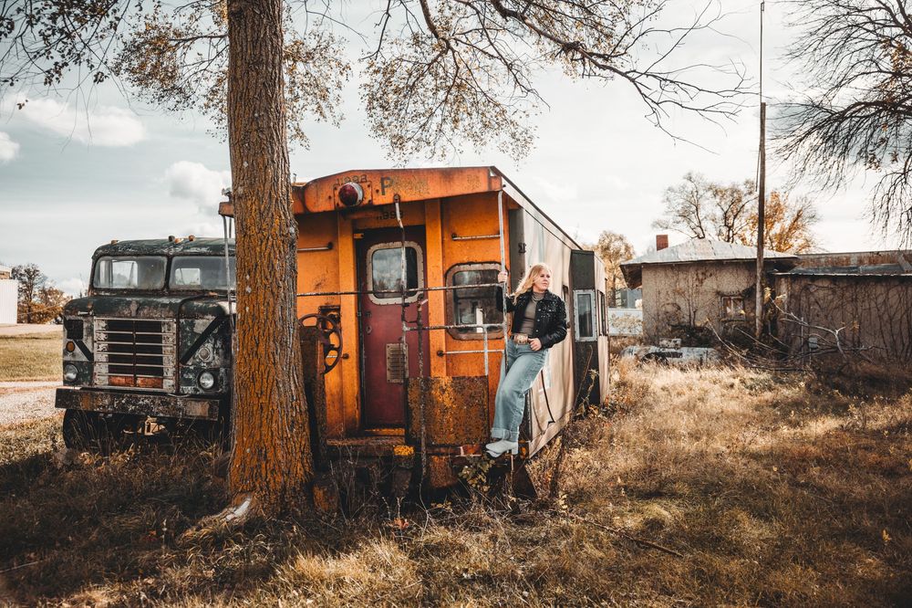 Rustic orange abandoned vehicles and buildings scattered among bare trees in a rural landscape at sunset.