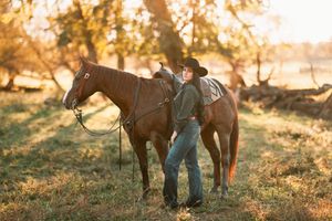 A cowboy stands with their horse in a golden field during sunset creating a warm silhouetted scene.