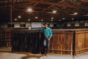A person in a teal shirt leans against a wooden stall inside a dimly lit barn or indoor riding arena.
