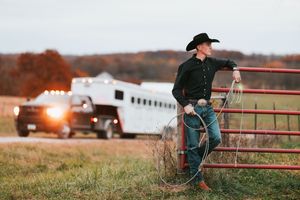 A cowboy sits on a red metal gate overlooking a rural farm field at sunset with barns in the background.