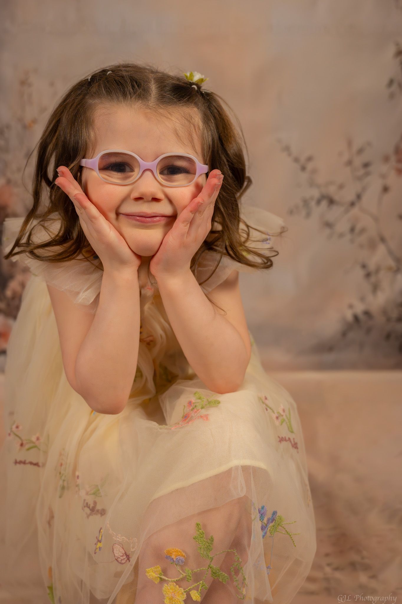 Adorable little girl in yellow tulle dress and purple glasses poses with hands on cheeks.