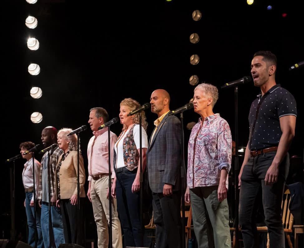 Group of diverse performers standing in a row on stage with microphones and dramatic stage lighting.