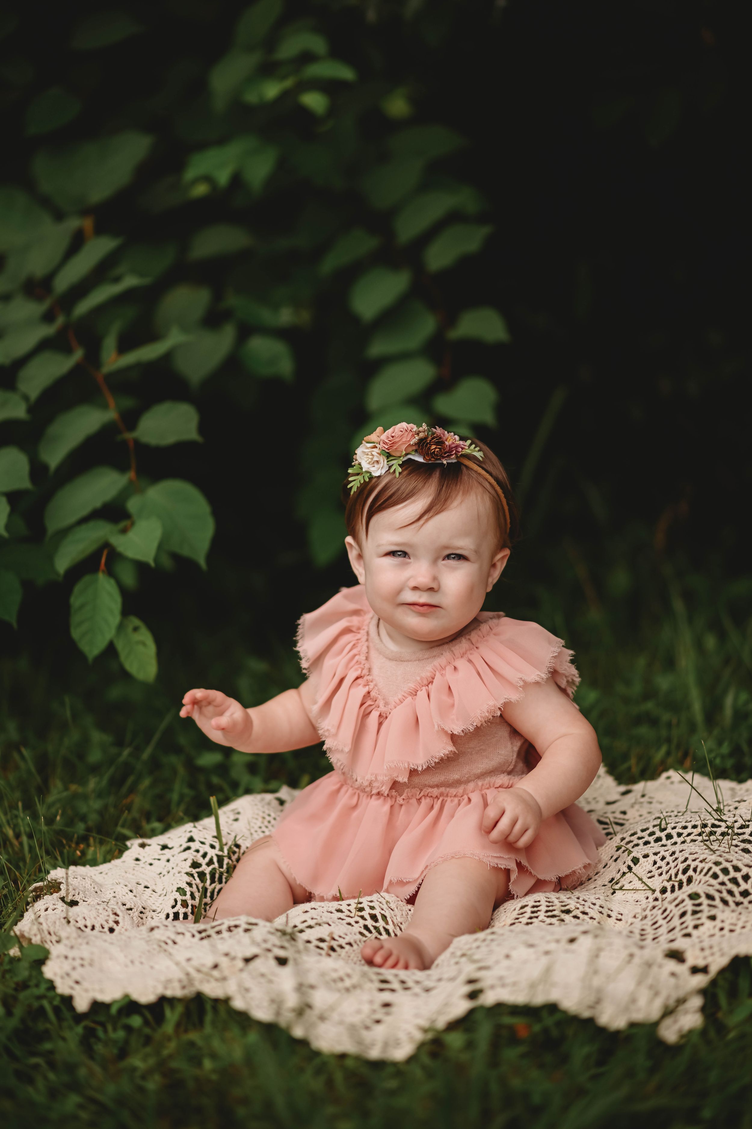 Baby wearing coral ruffle dress poses on cream lace blanket with greenery backdrop.