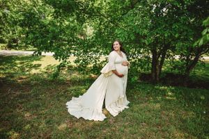 Artistic maternity photography with billowing cream dress against green forest backdrop.